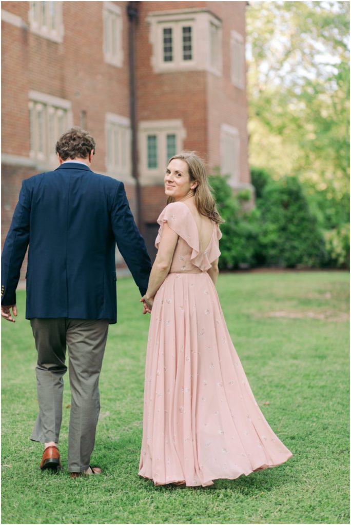 A woman in a blush pink dress holds hands with her fiance and smiles at the camera
