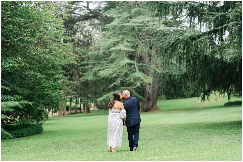 A girl in a dress and a man in suit kiss under trees at their summer engagement session