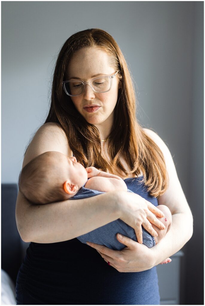 Mother with long red hair and glasses gently holding her newborn baby in a softly lit room during an in-home Richmond newborn photography session.